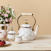 MacKenzie Childs | Rosy Check Tea Kettle, 2 QT, with pink-and-white checkerboard design, brass handle, and glass finial, elegantly displayed with matching accessories and flowers.