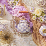 MacKenzie-Childs | Violet Check Tea Kettle, 2 Qt, with wooden handle, surrounded by matching checkered ceramics and spring blooms, on a decorative surface.