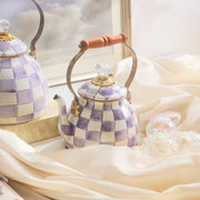 MacKenzie-Childs Violet Check Tea Kettle, 2 Qt, featuring a lavender checkerboard pattern, brass fittings, and a wood handle, elegantly displayed on ivory fabric.
