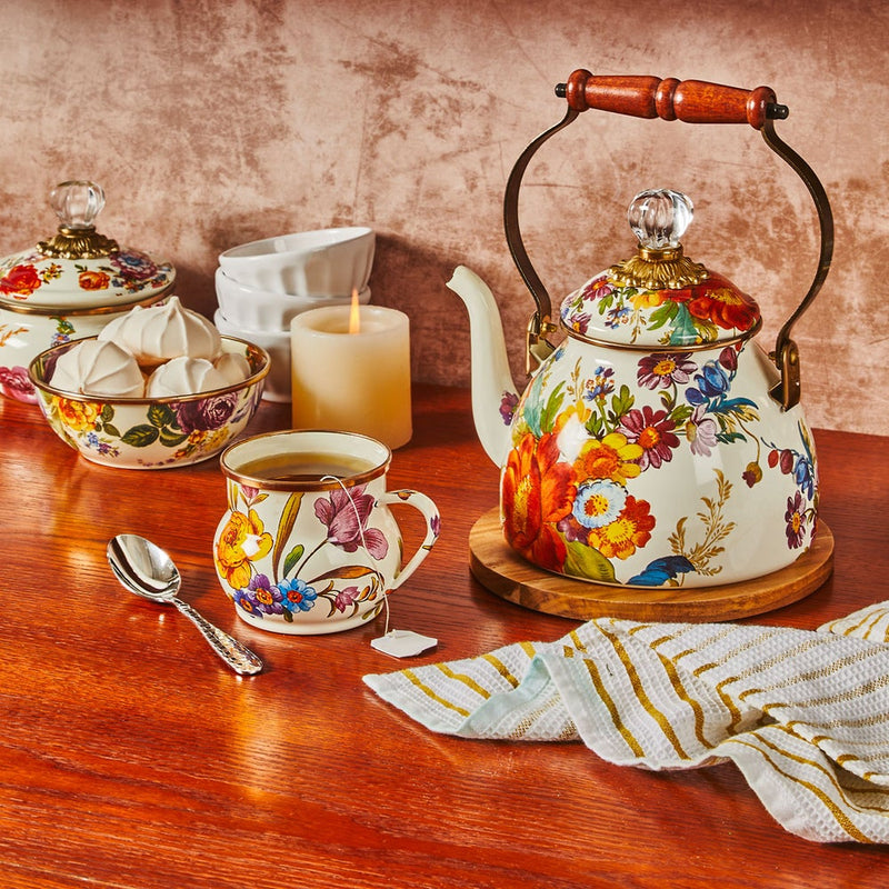 MacKenzie Childs White Flower Market Tea Kettle with wooden handle, on trivet, surrounded by matching tea cup, confections bowl, and lit candle on a cozy table.