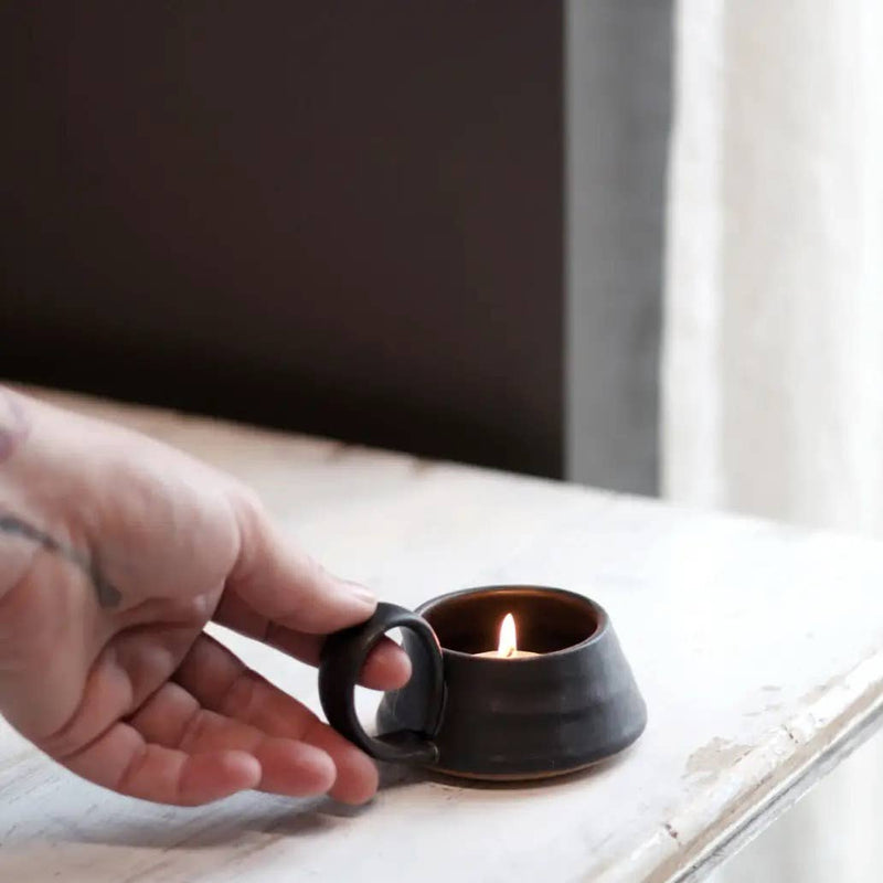 Gravesco Pottery - Tea Lite Candle Holder in Black with a lit candle, featuring a ring handle, sitting on a wooden surface, perfect for portable candlelight.