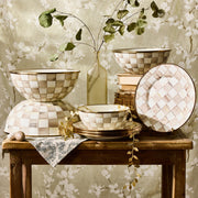 MacKenzie-Childs Mocha Check Small Everyday Bowl on rustic table with ceramic set, vintage books, glass vase, blue floral napkin, gold sprig, and decorative eggs.