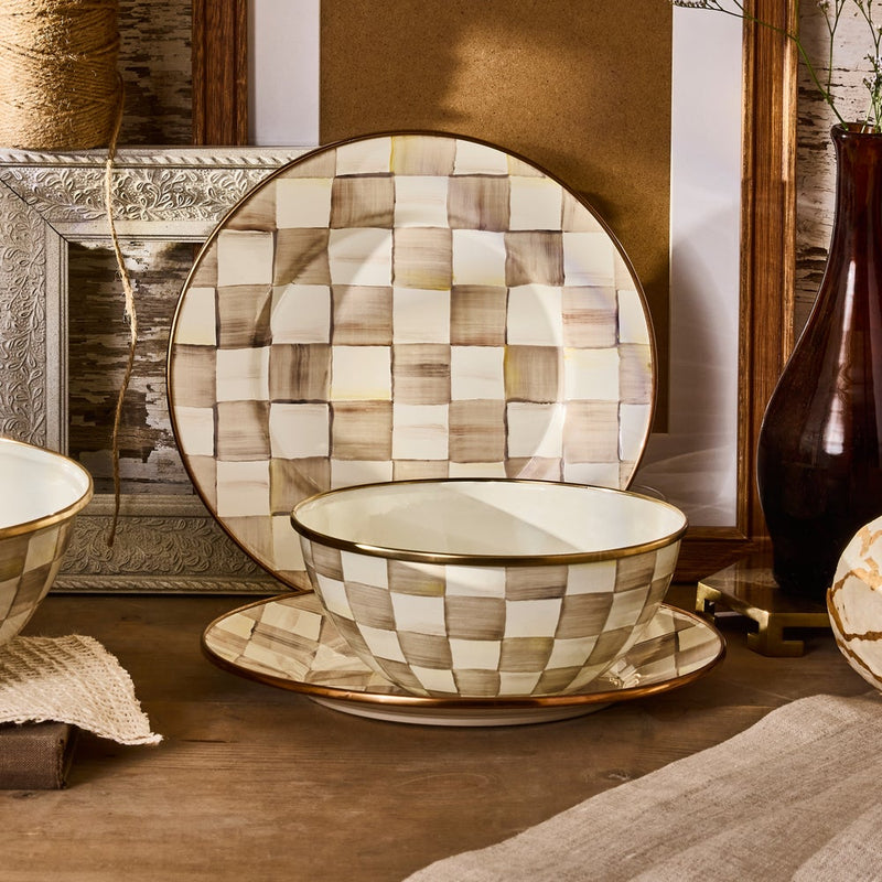 Mocha Check Small Everyday Bowl by MacKenzie Childs, hand-painted with coffee checks, sits on a plate with rustic decor, embodying warm, rustic-chic style.