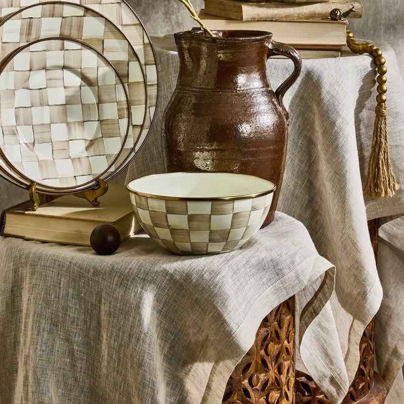 MacKenzie Childs | Mocha Check Small Everyday Bowl displayed in a rustic setting with matching ceramics, an earthenware jug, and vintage books on a wooden stool.
