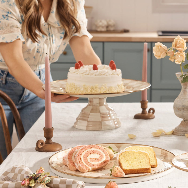 MacKenzie Childs | Mocha Check Large Pedestal Platter elegantly displays a white-iced cake, surrounded by sliced cakes and strawberries, set on a beautifully arranged tabletop.