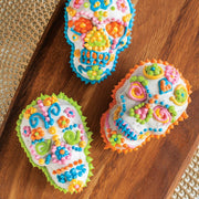 Nordic Ware | Skull Cakelet Pans - Skull Bites: Three intricately decorated skull-shaped cupcakes, showcasing vibrant Day-of-the-Dead designs on a wooden platter, ideal for creative Halloween baking.