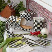 MacKenzie Childs | Courtly Check Utensil Holder with wooden spoons, surrounded by checkered kitchen items, knives, and fresh produce on a marble countertop.