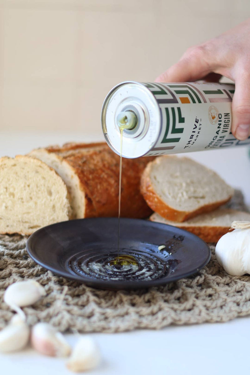 Pouring olive oil into Gravesco Pottery - Garlic Grater Plate in Black, surrounded by garlic cloves and bread, showcasing its use for flavorful dips.