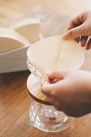 A person inserts a filter into a glass-and-wood dripper, highlighting HARIO USA - V60 Paper Filters: White / 100 Sheets Boxed, enhancing pour-over brewing.