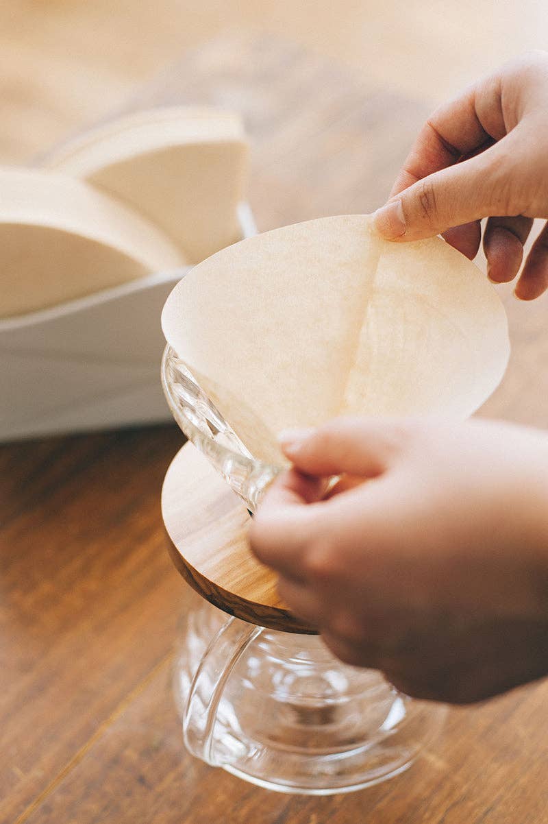 A person inserts a filter into a glass-and-wood dripper, highlighting HARIO USA - V60 Paper Filters: White / 100 Sheets Boxed, enhancing pour-over brewing.