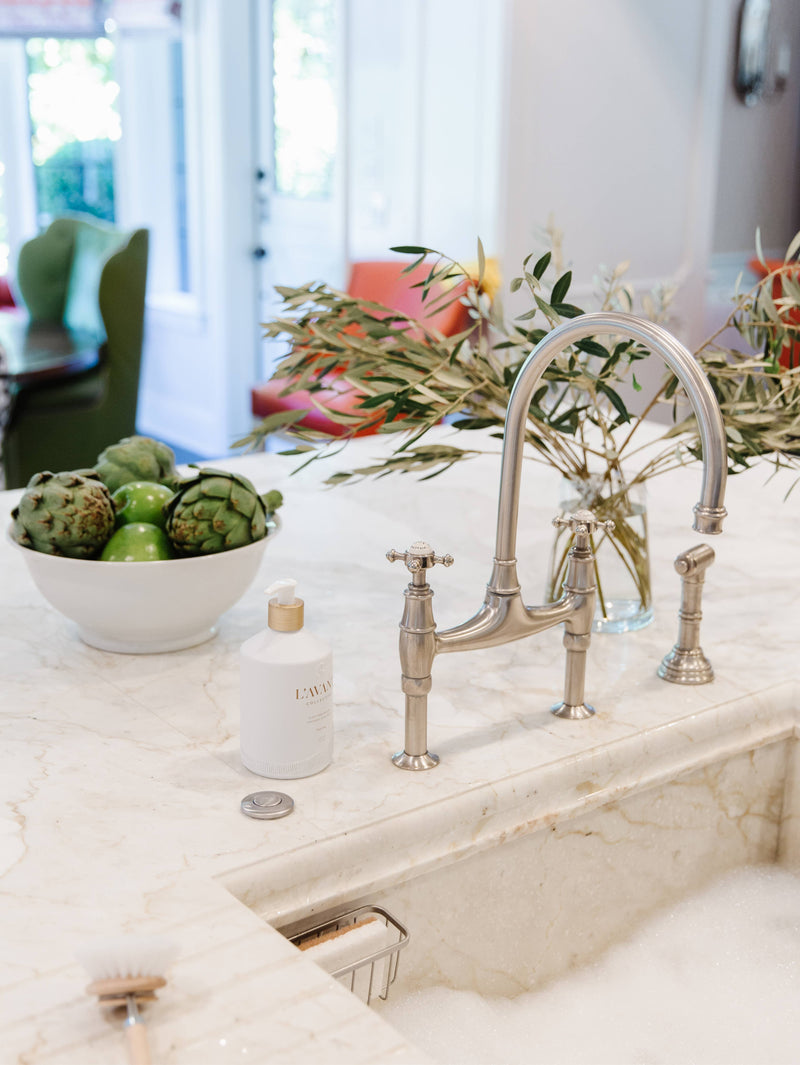 L'AVANT Collective - Fresh Linen High Performing Dish Soap in a matte white bottle beside a sink with fruit, enhancing kitchen elegance.