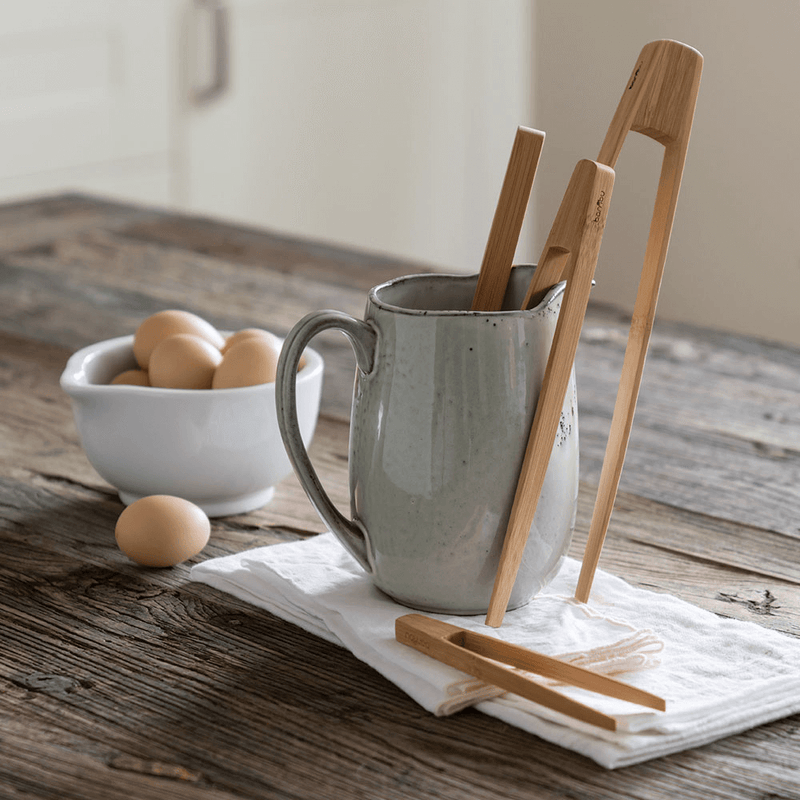bambu® - Small Bamboo Tongs on a rustic kitchen table, displayed with brown eggs and a ceramic pitcher, capturing a warm, natural kitchen aesthetic.