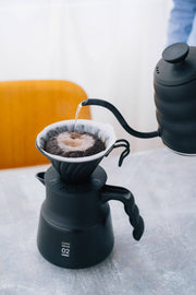 HARIO USA - V60 Metal Dripper: Silver in action with a gooseneck kettle, showcasing precise pour-over brewing on a black server carafe.