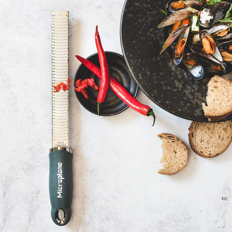 Microplane Premium Zester, Grater - After Dark Grey, showcasing its blade with grated red pepper next to mussels, highlighting its efficiency in zesting and grating.