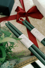 Mo&Co Home - Forest Green Dipped Beeswax Taper Candles rest on a festively wrapped gift, surrounded by evergreen sprigs, highlighting their holiday-ready elegance.
