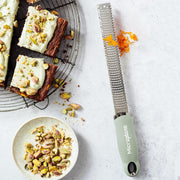 Microplane | Premium Zester, Grater - Sage Green on countertop with orange zest, alongside pistachio-topped blondies and pistachios, ideal for zesting and grating tasks.