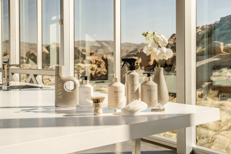 L’AVANT Collective Multipurpose Cleaner Ambre Santal on a modern countertop with neutral bottles, scrub brush, and orchid; overlooks a desert landscape.