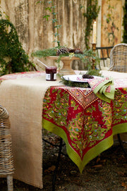 Rustic holiday table with Couleur Nature - Noel Red & Green French Tablecloth, featuring festive decor, fir sprigs, and a pine cone accent, set in a garden ambiance.