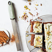 Microplane Premium Zester, Grater - Sage Green shown grating spice beside pistachio-frosted cake squares on a cooling rack, exemplifying its versatility in baking.