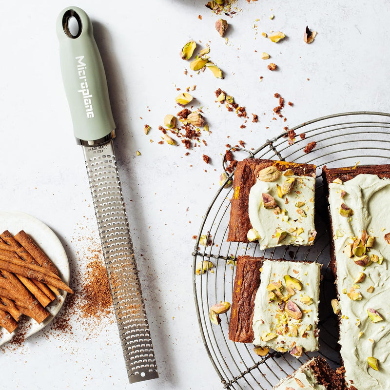 Microplane Premium Zester, Grater - Sage Green shown grating spice beside pistachio-frosted cake squares on a cooling rack, exemplifying its versatility in baking.