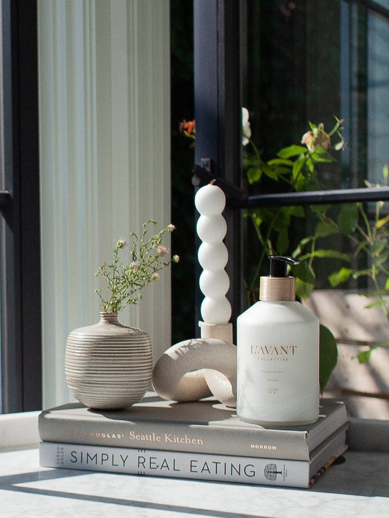 L'AVANT Collective - Fresh Linen Hand Lotion displayed among elegant vases, emphasizing its luxurious glass bottle design and premium skincare qualities.