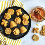 Nordic Ware Busy Bee Bitelet Pan featuring 16 cavities for creating bite-sized treats, shown with a plate of cookies and a honey dipper jar nearby.
