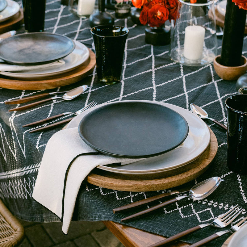 ATELIER SAUCIER - Cream Burlap Noir Napkins elegantly drape a stylish place-setting, enhancing the rustic charm with premium reclaimed fabric and sleek black edging.