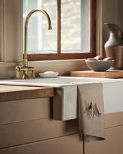 Coral & Tusk - Christmas Cardinals Tea Towel draped over a farmhouse sink, featuring embroidered cardinals on evergreen sprigs, complementing the rustic kitchen setting.