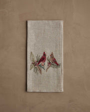 Coral & Tusk - Christmas Cardinals Tea Towel with embroidered cardinals on holly and pine branches, folded on a taupe surface, ideal for holiday gifting.