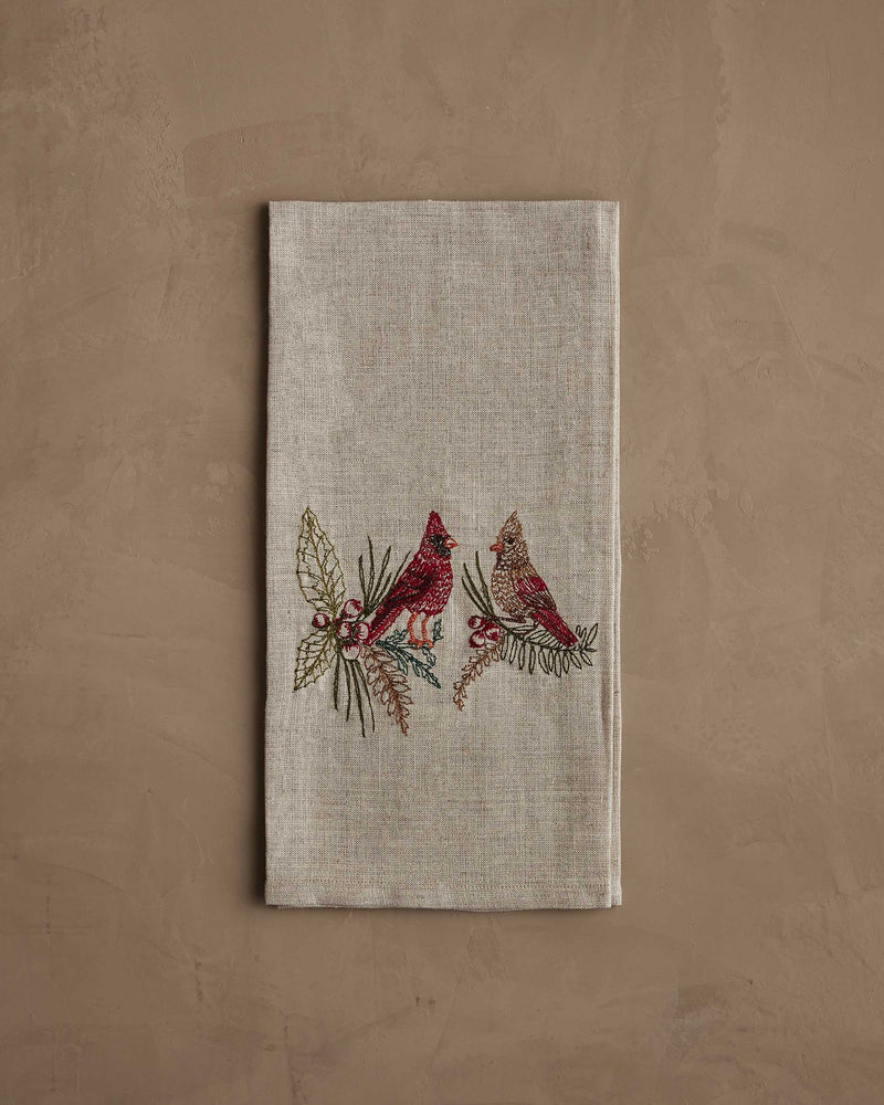 Coral & Tusk - Christmas Cardinals Tea Towel with embroidered cardinals on holly and pine branches, folded on a taupe surface, ideal for holiday gifting.