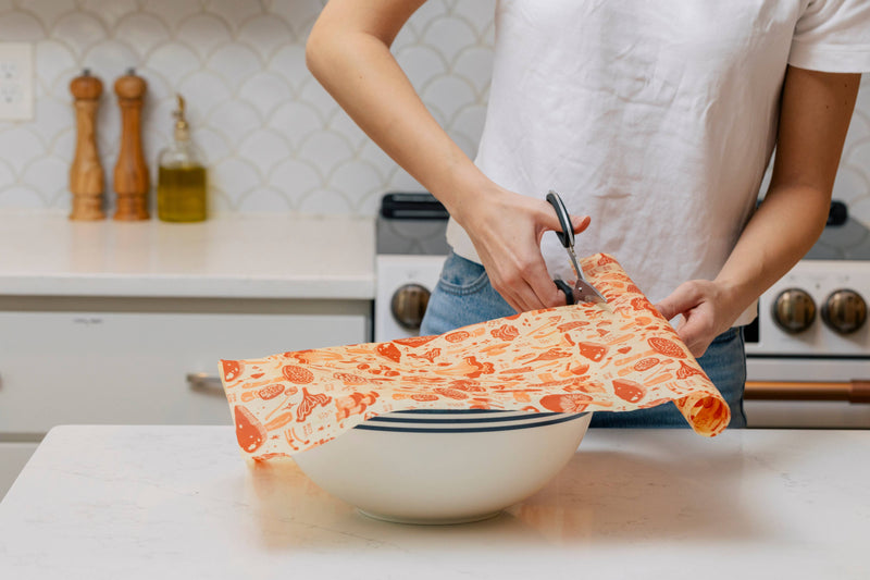 Woman cuts Bee's Wrap - Cut-to-Size Roll - Mushroom Magic in kitchen, preparing to cover a mixing bowl. Salt, pepper, and olive oil visible nearby.