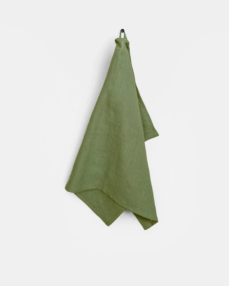 Green linen tea towel with hanging loop, made from European flax, displayed on a hook. Durable and absorbent, ideal for kitchen use.
