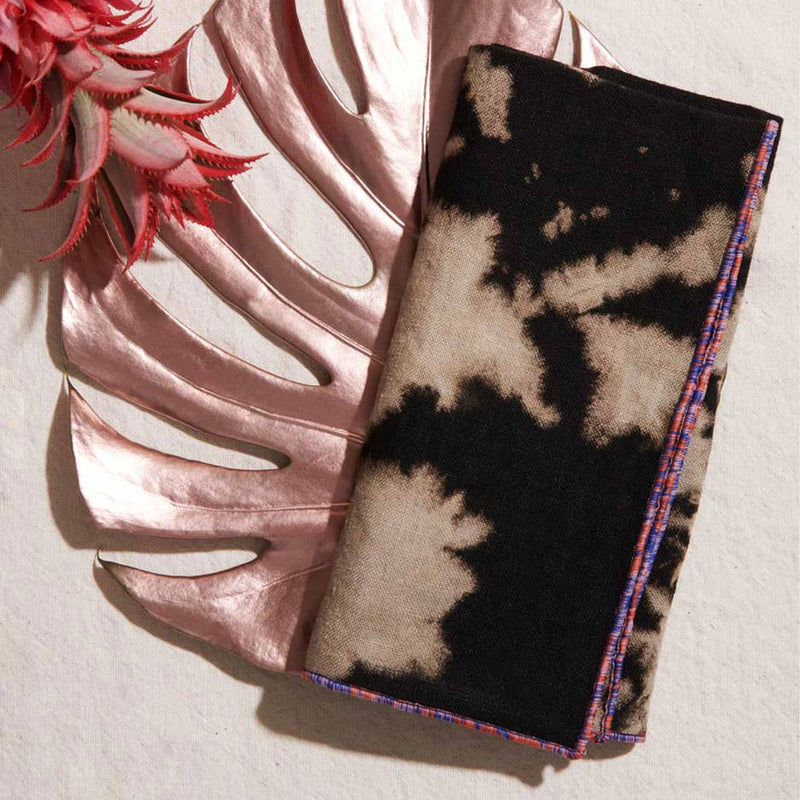 ATELIER SAUCIER - Sundown Linen Napkins in a tie-dye pattern, set of 4, displayed on a rose-gold monstera leaf with a red-pink succulent accent.