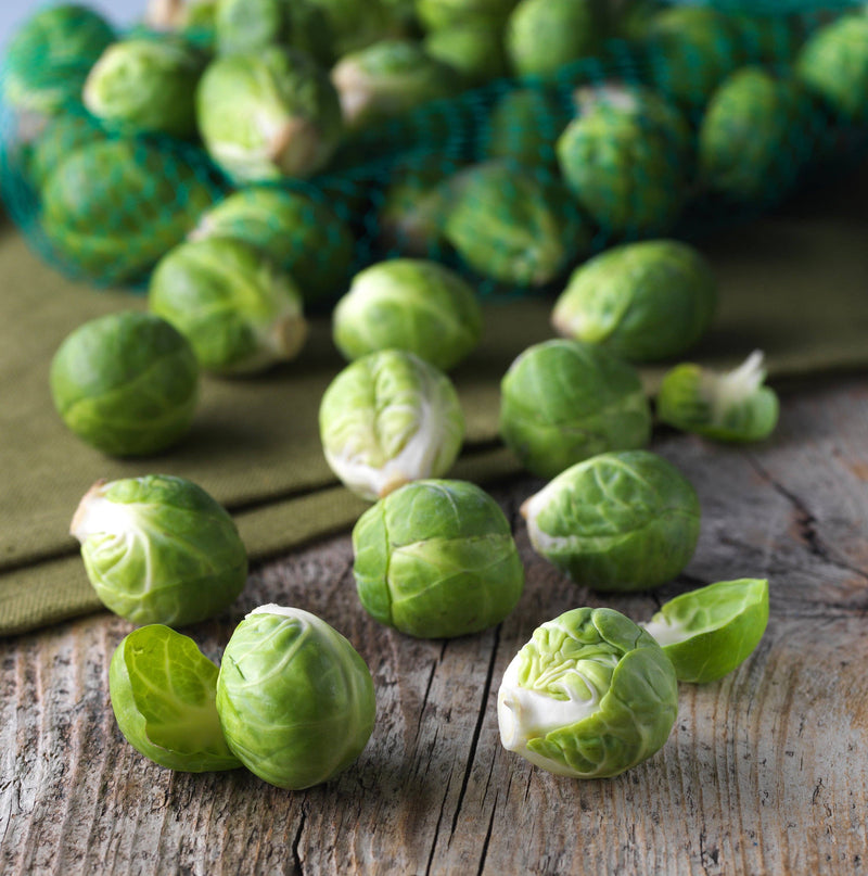 Bentley Seed Co. - Brussel Sprouts-Long Island 1890's seeds with fresh Brussels sprouts scattered on a wooden surface, mesh produce bag in background.