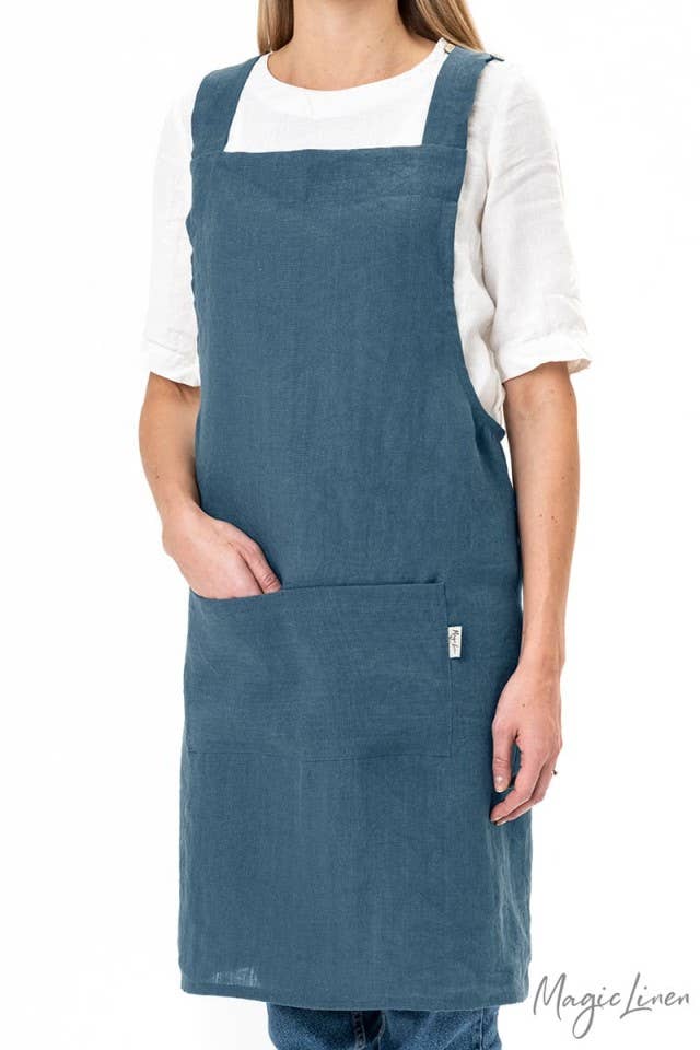 Woman wearing MagicLinen's Pinafore cross-back linen apron with twin-sectioned front pocket, ideal for cooking or gardening. Available in various colors, unisex design.