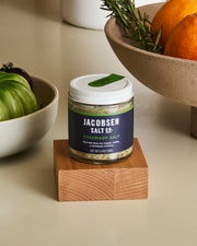 Jacobsen Salt Co. Rosemary Salt in a 3.5 oz glass jar on wood block, with bowls of green tomatoes and oranges, perfect for enhancing roasted lamb and focaccia.