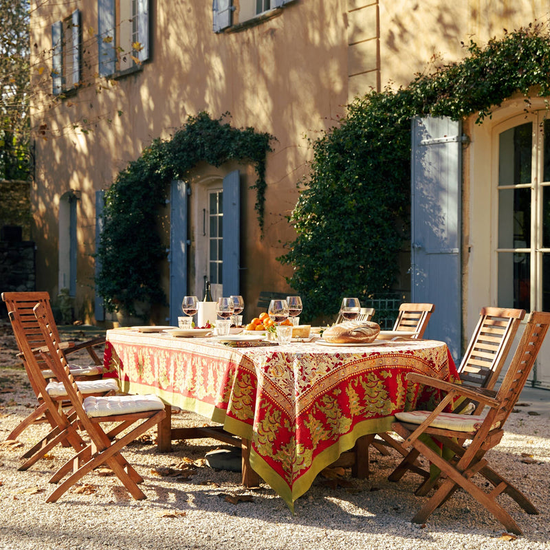 French tablecloth by Couleur Nature, Noel Red & Green, adorns a wooden table with wine, bread, and fruit, set for an al fresco meal.
