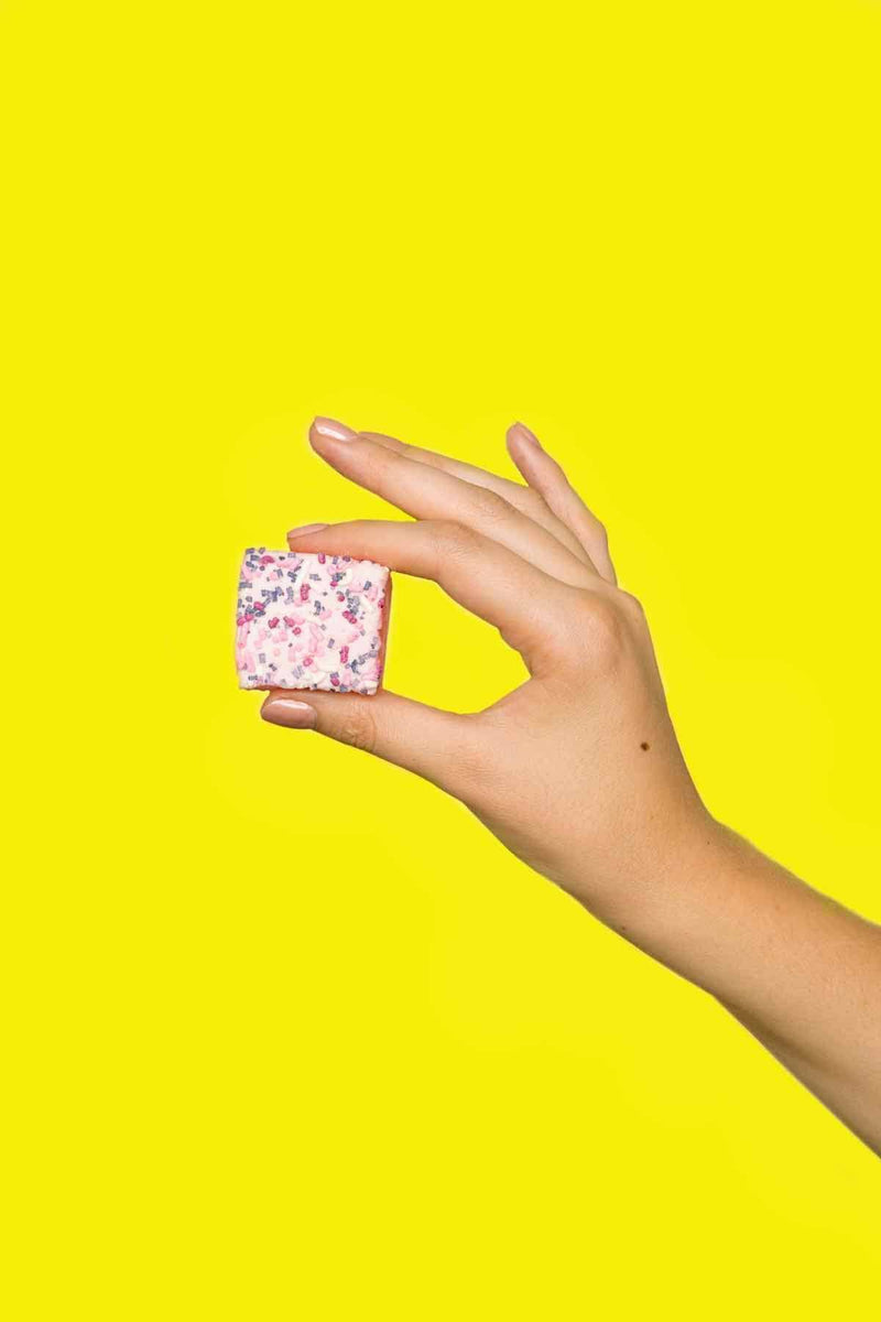 Malvi Mallow - Cotton Candy 4-Pack Marshmallows, Case of 12 Packs: A hand holds a pink marshmallow cube with sprinkles, showcasing its airy texture against a bright yellow background.