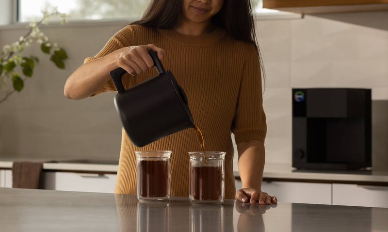 Woman using FELLOW - Aiden Precision Coffee Maker: Matte Black to pour coffee into two glasses, highlighting its precision and ease of use.