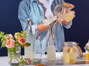 Home-brewing scene with Le Parfait 500ml French Glass Swing Top Bottle receiving strained, pale-yellow fermented liquid, surrounded by jars, utensils, and decorative Mason jar with roses.