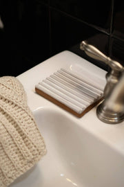 Gravesco Pottery - Soap Dish | White: Close-up of a handmade ceramic soap dish on a wooden base, showcasing its unique ridged design beside a satin-nickel faucet in a bathroom setting.