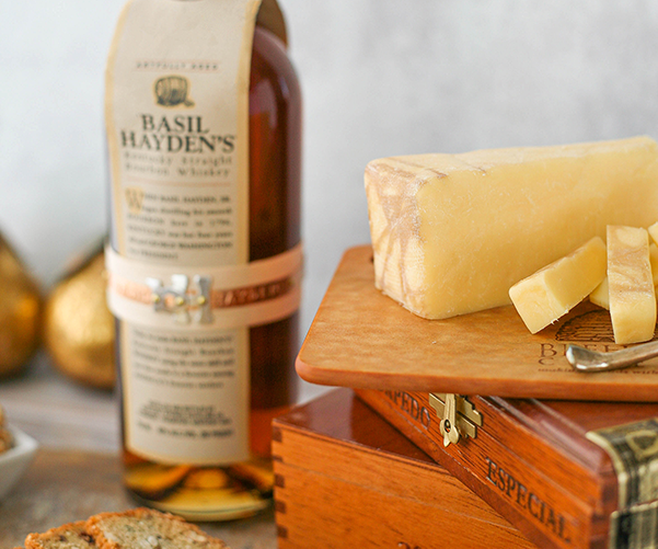 Beehive Cheese Co. LLC | Pour Me A Slice - 4 oz Wedge, close-up of cheddar infused with Basil Hayden Bourbon, displayed on a wooden surface.