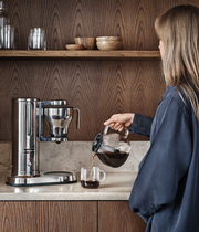 A woman pours coffee into an Aarke Glass Carafe Coffee Maker's replacement glass carafe, part of the Athens Cooks' curated kitchen collection.