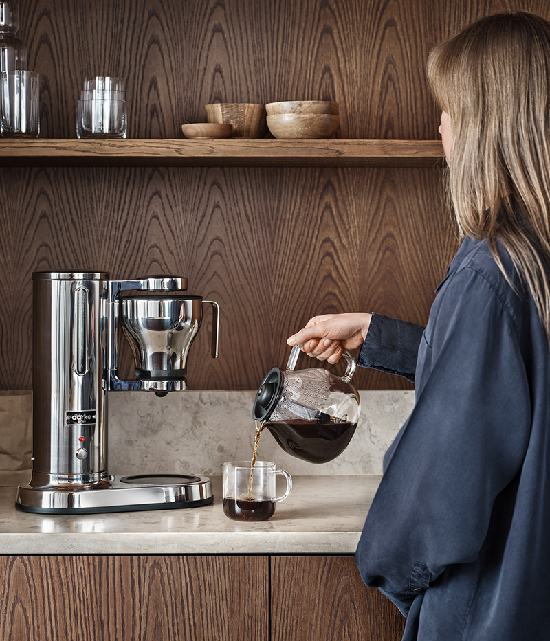 A woman pours coffee into an Aarke Glass Carafe Coffee Maker's replacement glass carafe, part of the Athens Cooks' curated kitchen collection.