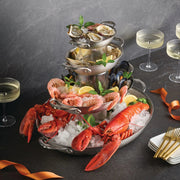 Hestan Provisions Large Clad Oval Roaster elegantly presents a three-tier seafood platter with oysters, clams, lobster, and prawns, surrounded by champagne glasses and dining utensils.