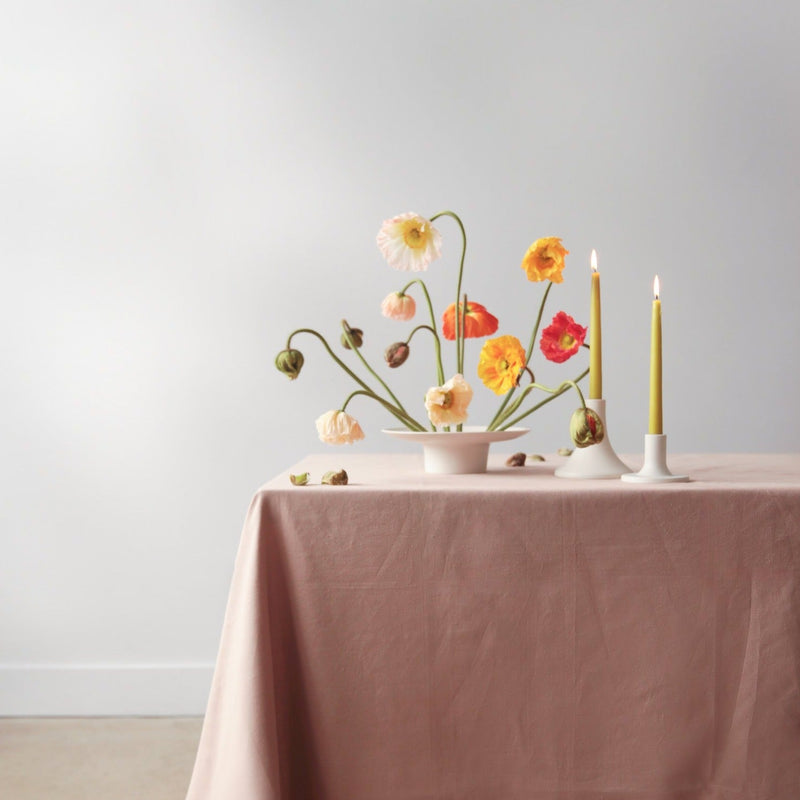 The Floral Society | Candle Holders, Grand, Matte White Tall elegantly display mustard-yellow taper candles, complementing a modern floral arrangement on a draped table.
