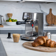 Breville | Bambino Plus espresso machine on a marble countertop with latte art, tamper, milk pitcher, and croissants, showcasing professional-grade home coffee-making essentials.