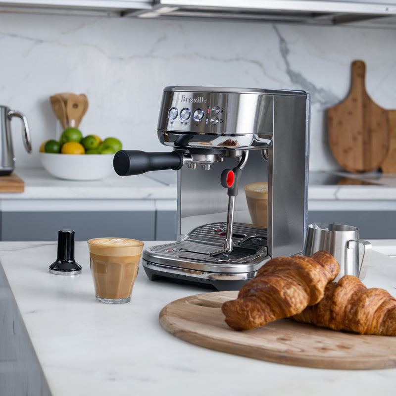 Breville | Bambino Plus espresso machine on a marble countertop with latte art, tamper, milk pitcher, and croissants, showcasing professional-grade home coffee-making essentials.