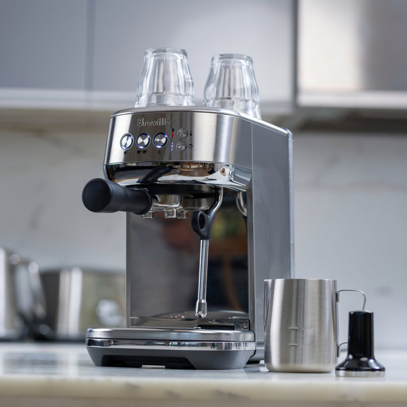 Breville | Bambino Plus espresso machine on counter with portafilter handle, glass cups on top, steam wand, and milk-frothing pitcher nearby, ideal for precise home brewing.