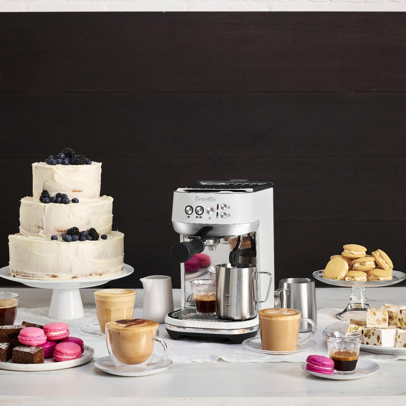 Breville | Bambino Plus espresso machine on display with coffee drinks, pastries, and a tiered cake, highlighting its professional-grade brewing capabilities.
